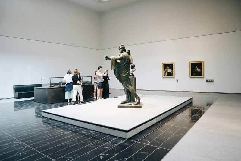 Salle d'exposition du Louvre Abu Dhabi avec une statue antique au centre et des visiteurs observant les œuvres