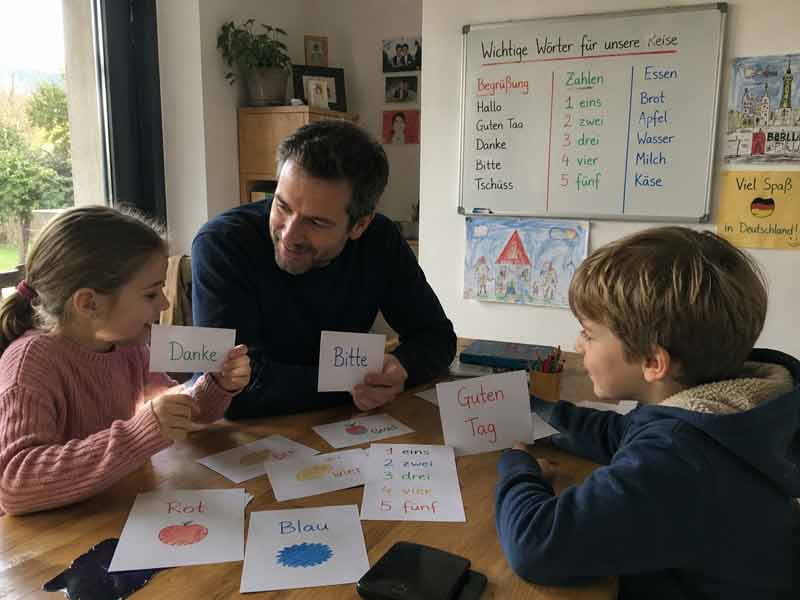 Père et enfants apprenant les premiers mots d'allemand en famille avec des flashcards avant le départ en voyage