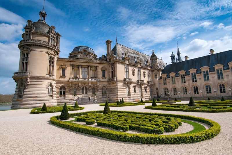 Jardins à la française du château de Chantilly avec sa façade Renaissance et les Grandes Écuries en arrière-plan, sous un ciel bleu nuageux