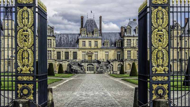 Entrée du château de Fontainebleau encadrée par une grille ornée de dorures, avec vue sur l'escalier en fer-à-cheval et la cour d'honneur
