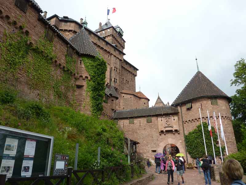 Entrée du château du Haut-Koenigsbourg en Alsace avec ses murailles en grès rose, ses tours médiévales et des visiteurs sous un ciel couvert