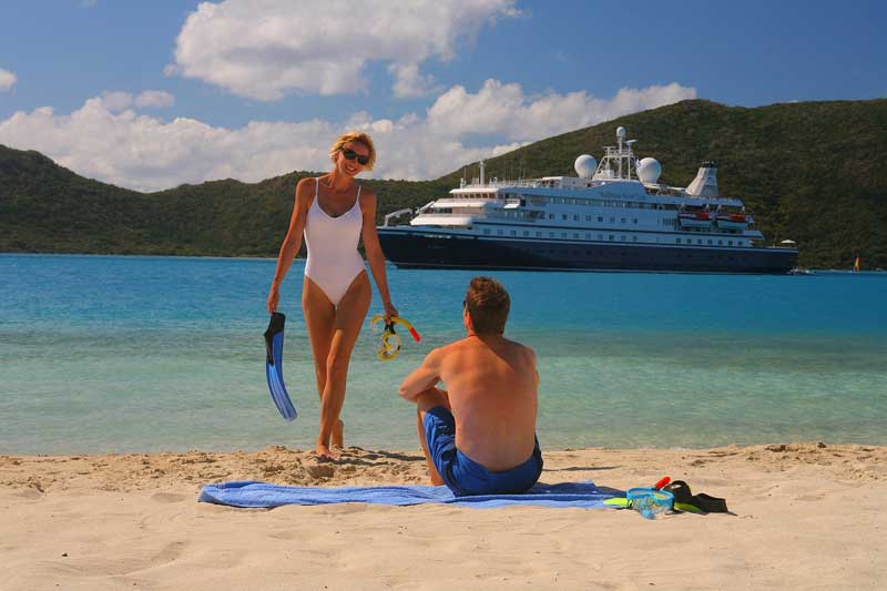 Photo d'une jeune femme en maillot de bain une pièce qui sort de la mer avec des palmes à la main, on voit un bateau de
croisière pour célibataires en arrière plan