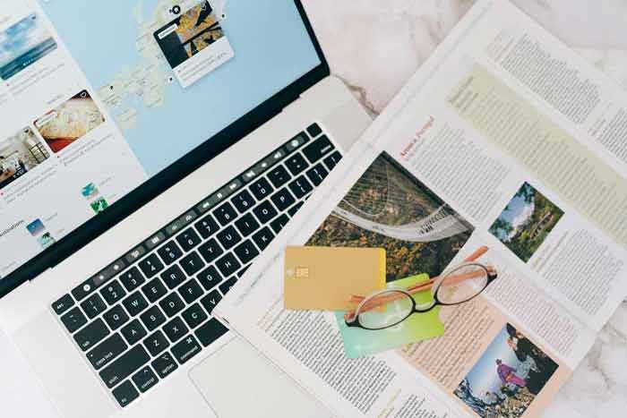 Ordinateur portable affichant une carte, avec un magazine de voyage, des cartes bancaires et des lunettes, illustrant la recherche d'informations et la prparation financire pour un long voyage