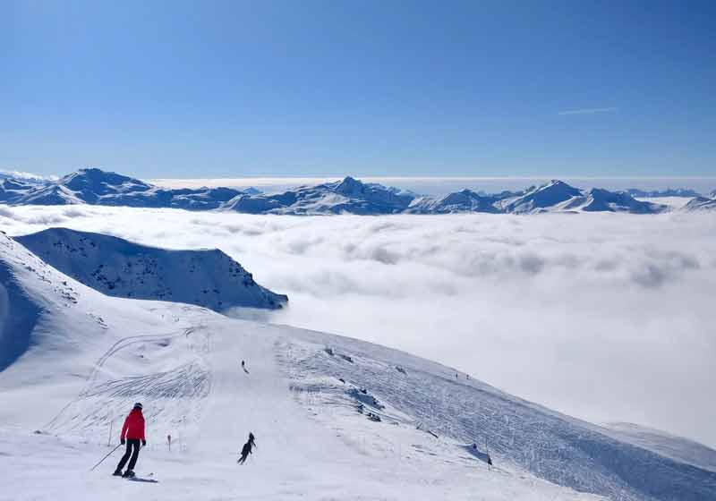 Skieurs sur une piste enneigée des Alpes au-dessus d’une mer de nuages, sous un grand ciel bleu, pour un week-end au ski en décembre