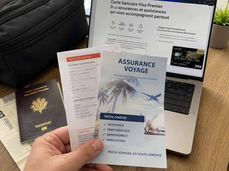 Brochure d'assurance voyage tenue à la main devant un passeport français et un écran affichant les garanties d'une carte bancaire 
							Visa Premier