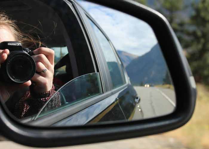 Reflet dans le rtroviseur d'une voiture montrant une personne prenant une 
						photo avec un appareil photo Canon, sur une route borde de montagnes et de forts
