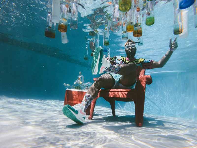 Homme assis sous l'eau dans une piscine pendant le spring break, entouré de bouteilles flottantes, portant des lunettes de soleil
et des vêtements colorés