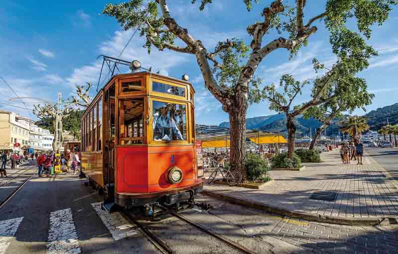 Ancien tramway en bois circulant sur le front de mer de Port de Sóller à Majorque, sous un ciel bleu et entouré de terrasses
animées et de montagnes