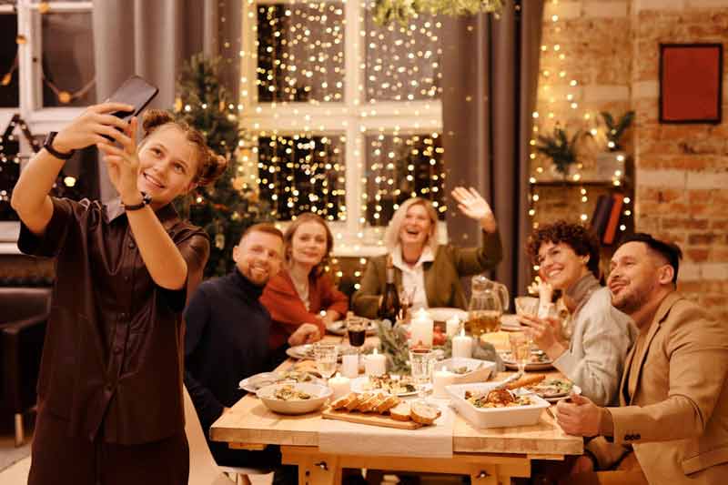 famille partageant un dîner de Noel autour d'une table, avec une ambiance chaleureuse et des guirlandes lumineuses en arrière-plan.
Une jeune femme prend un selfie pendant que les autres sourient.