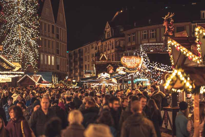 Marché de Noël de Francfort en soirée avec une foule de visiteurs, des stands illuminés, un grand sapin décoré et des bâtiments
traditionnels en arrière-plan. L'ambiance festive est accentuée par les lumières scintillantes et les décorations de Noël.