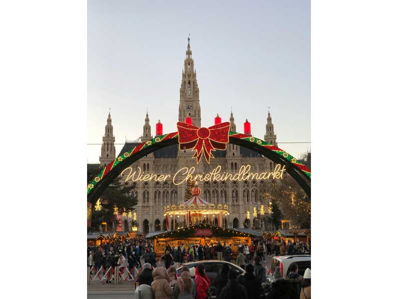 Marché de Noël de Vienne avec une arche lumineuse décorée de rubans rouges et verts portant l'inscription 'Wiener Christkindlmarkt',
devant le Rathaus, bâtiment emblématique de la ville. Une foule de visiteurs profite de l'ambiance festive au coucher du soleil.