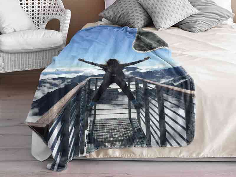 couverture personnalisée posée sur un lit, représentant une personne debout les bras écartés sur une passerelle
face à un panorama de montagnes et de nuages. L'ambiance est apaisante et inspirante, idéale pour un cadeau original.