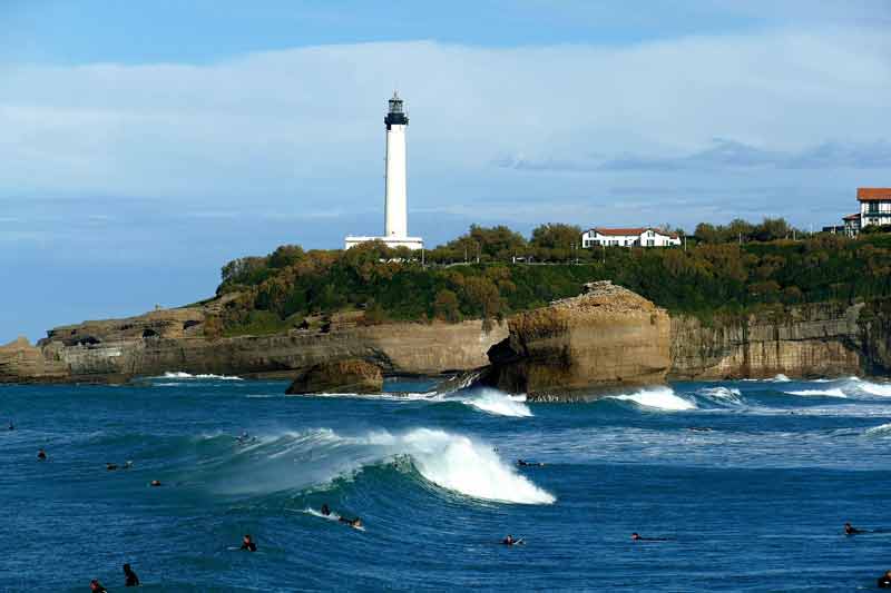 Surfeurs dans les vagues de l'Atlantique face au phare de Biarritz perché sur ses falaises rocheuses, Pays Basque, France