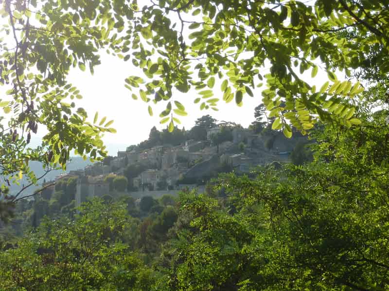 Village perché du Luberon entrevu à travers un cadre de feuillages verts, accroché à son éperon rocheux dans la lumière provençale, 
							Luberon, Provence