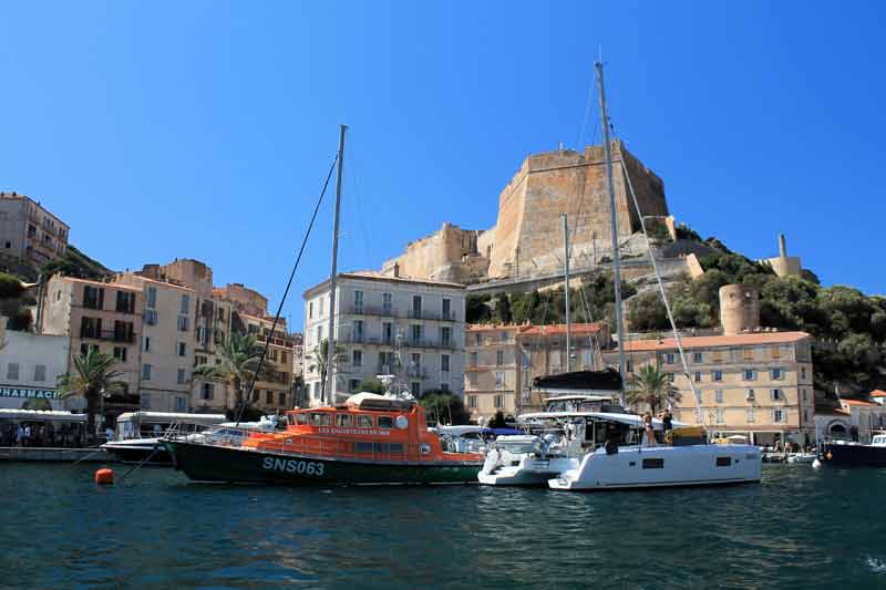 Vue du port de Bonifacio en Corse, avec des bateaux amarrés au premier plan et la citadelle perchée sur une falaise en arrière-plan sous un ciel bleu