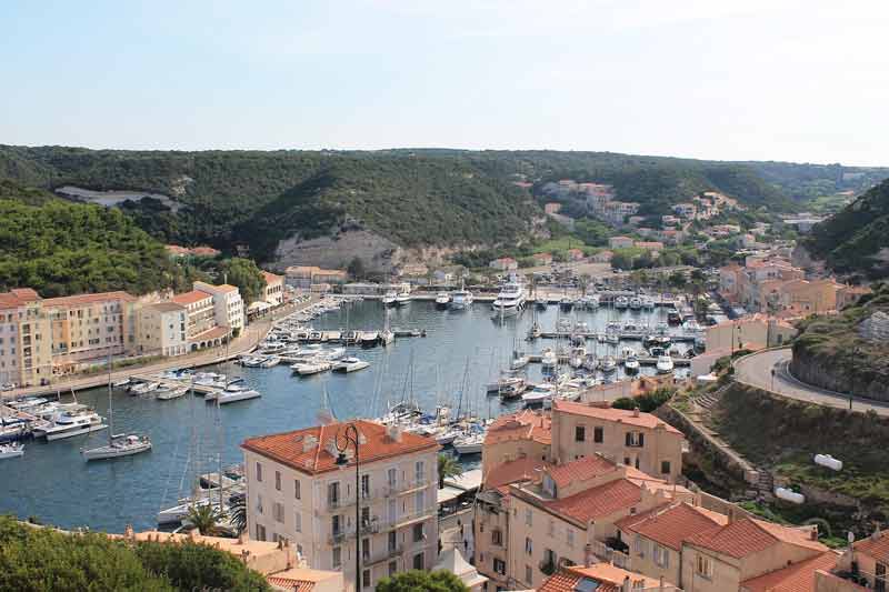 Vue panoramique sur le port de Bonifacio en Corse, entouré de collines verdoyantes et de bâtiments corses