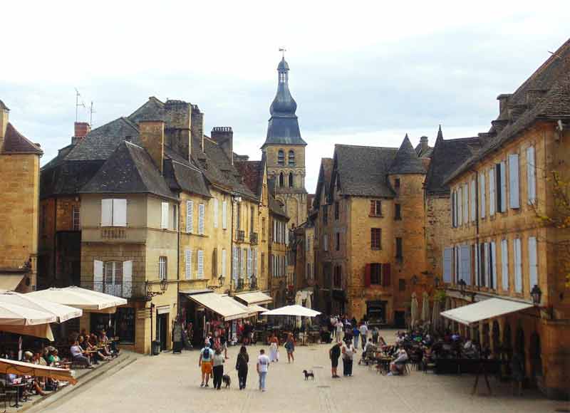 Place du Peyrou à Sarlat-la-Canéda en été avec ses terrasses de café, ses façades en pierre blonde et le clocher de la cathédrale