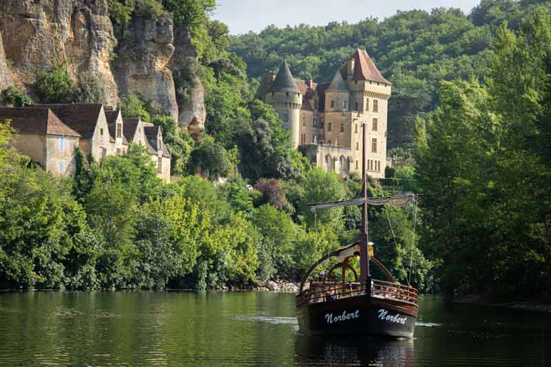 Gabarre traditionnelle naviguant sur la Dordogne au pied du château de la Malartrie et des maisons troglodytiques de La Roque-Gageac