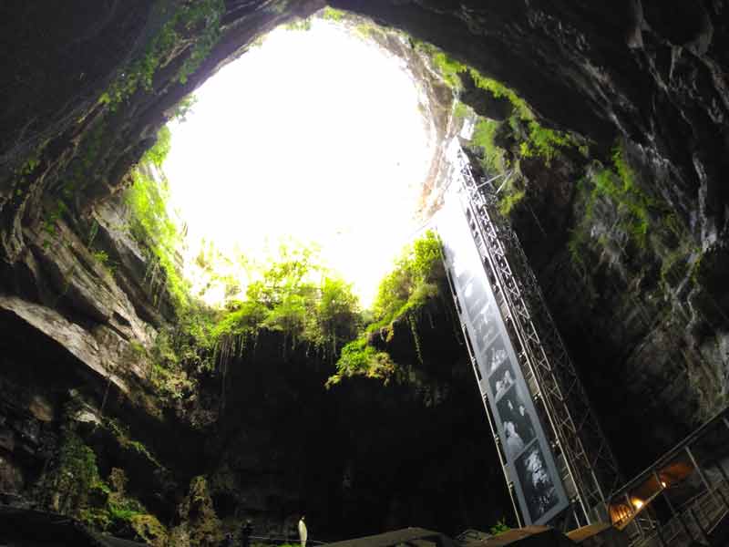 Vue depuis le fond du gouffre de Padirac vers l’ouverture baignée de lumière et de végétation, avec l’ascenseur métallique qui descend sous terre