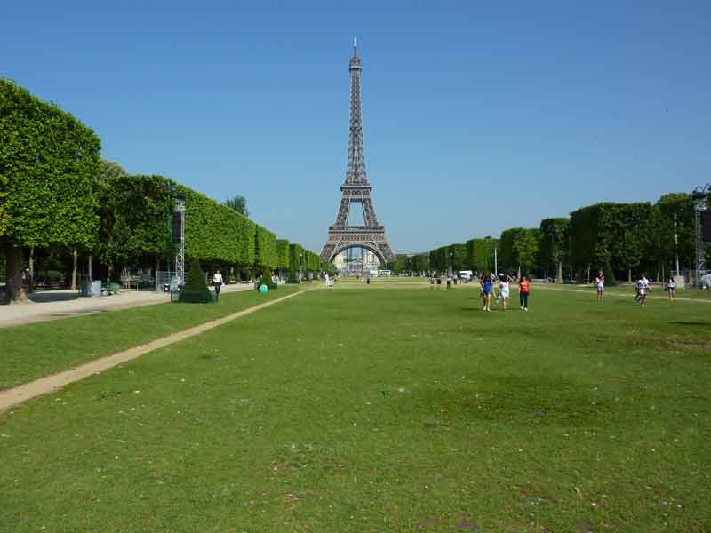 Vue de la tour Eiffel depuis le Champ-de-Mars, avec les pelouses et les allées bordées d’arbres au premier plan
