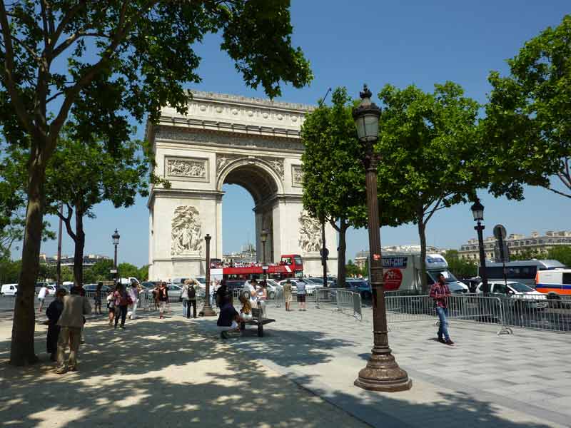 Arc de Triomphe à Paris vu depuis une allée arborée, avec des visiteurs et l’animation de la place en arrière-plan