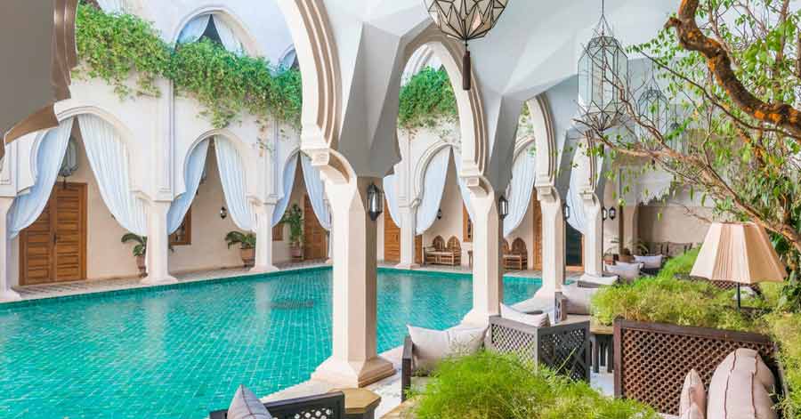 Piscine turquoise entourée d'arcades mauresques blanches dans un riad de luxe à Marrakech, avec lanternes marocaines suspendues et végétation grimpante