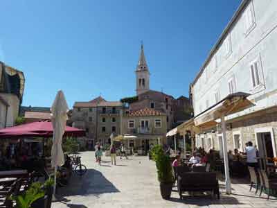 Place animée bordée de cafés et maisons en pierre avec le clocher d’une église en arrière-plan, sous un ciel 
												bleu à Hvar, en Croatie