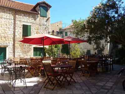 Terrasse ensoleillée d’un café avec parasols rouges et chaises en bois, dans une cour pavée entourée de 
												maisons en pierre à Hvar, en Croatie