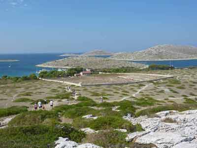 Paysage aride du parc national des Kornati en Croatie, avec des randonneurs marchant vers une bâtisse entourée 
												de murets de pierre, face à la mer Adriatique