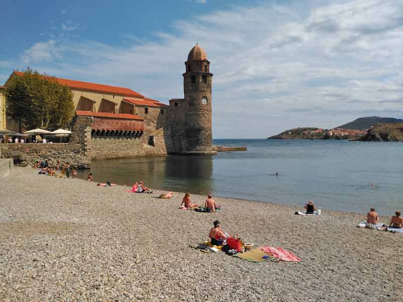Plage de galets à Collioure avec le clocher de l’église Notre-Dame-des-Anges, la mer calme et les baigneurs au premier plan