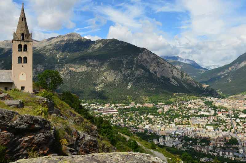 Vue panoramique sur Briançon depuis Puy-Saint-Pierre, avec l’église perchée au premier plan et les montagnes des Hautes-Alpes en toile de fond