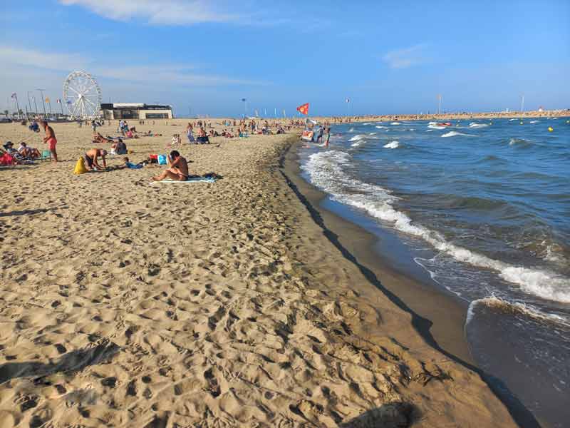 Plage animée de Canet-en-Roussillon avec baigneurs, sable doré, mer agitée et grande roue en arrière-plan sous un ciel estival