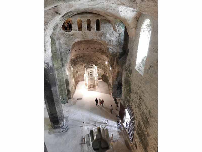 Vue intérieure de l'église souterraine monolithique d'Aubeterre-sur-Dronne avec son impressionnant espace creusé dans la roche, un baldaquin sculpté au centre et des visiteurs explorant le site (Charente, France)