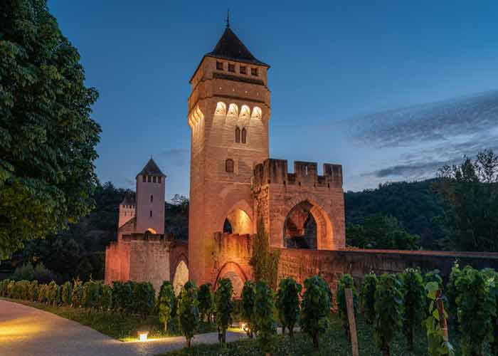 Pont Valentr  Cahors illumin au crpuscule, avec ses tours mdivales 
							en pierre et un sentier bord de vignes au premier plan