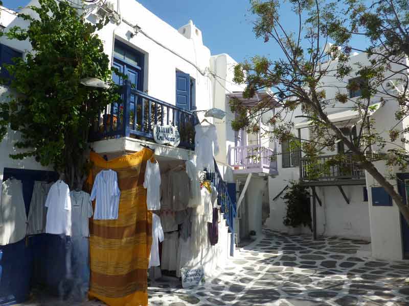 Boutique de vêtements dans une ruelle fleurie de Chora à Mykonos, avec maisons blanches et balcons colorés