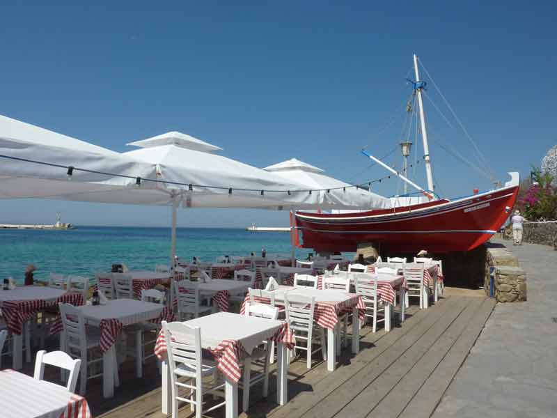Terrasse d'un restaurant en bord de mer à Little Venice à Mykonos avec nappe à carreaux rouges et bateau de pêche rouge en décoration