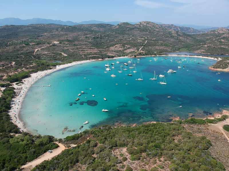 Vue aérienne de la plage de Rondinara en Corse, baie en forme de coquillage aux eaux turquoise avec bateaux au mouillage