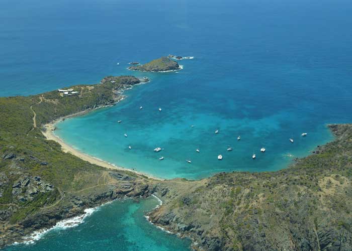 Vue aérienne d'une baie turquoise à Saint-Barthélemy, entourée
de collines rocheuses et arides, avec une plage de sable fin et plusieurs bateaux de plaisance ancrés dans des eaux cristallines