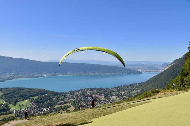 Parapentiste gonflant sa voile verte sur l’aire de décollage du col de la Forclaz, avec vue plongeante sur le lac d’Annecy