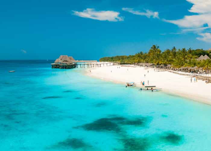 Plage paradisiaque de Zanzibar avec sable blanc, eau turquoise cristalline, petits bateaux traditionnels sur l'eau et un ponton en bois menant  des structures sur pilotis, borde de palmiers sous un ciel bleu clatant